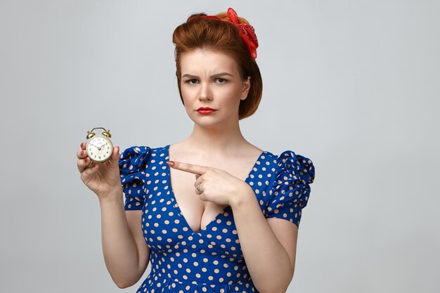 Studio shot of angry beautiful young woman dressed in vintage clothes looking at camera with annoyed expression, pointing index finger at alarm clock in her hand, meaning: You are late again