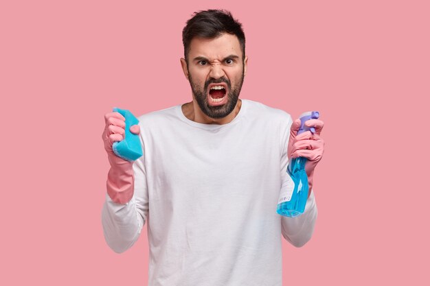 Studio shot of angry unshaven young man opens mouth from anger, holds spray and sponge, frowns face in discontent, expreses negative emotions
