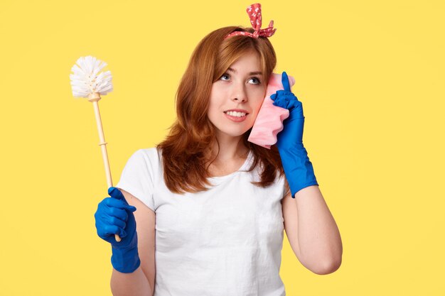 Studio shot of attentive housemaid holds brush, uses sponge as mobile phone, dicusses something with friend, dressed in casual clothes, poses on yellow. Cleaning and hygiene concept