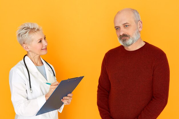 Studio shot of attractive confident middle aged female doctor with stethoscope aroudn her neck holding clipboard, handwriting symptoms while treating sad usnhaven senior man, ordering blood test