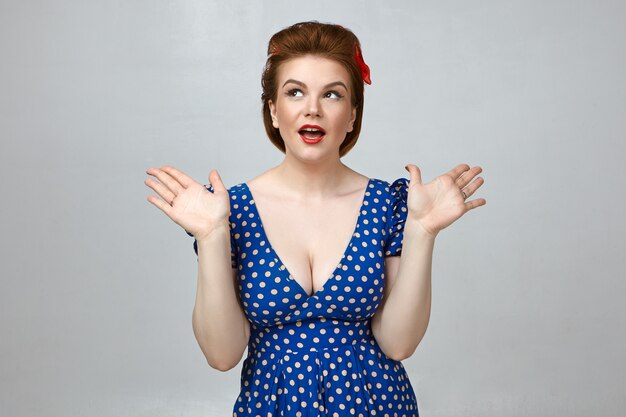 Studio shot of attractive gorgeous young woman wearing stylish retro dress with low cut and red lipstick gesturing emotionally, exclaiming in excitement