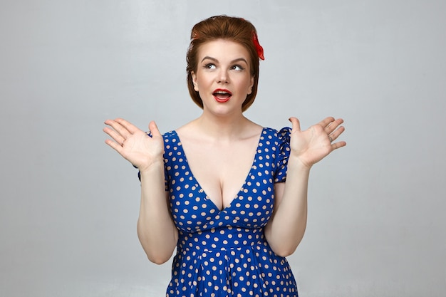 Studio shot of attractive gorgeous young woman wearing stylish retro dress with low cut and red lipstick gesturing emotionally, exclaiming in excitement