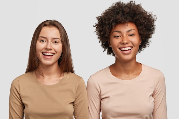 Studio shot of attractive joyful interracial best friend being in good mood, stand next to each other, smile broadly