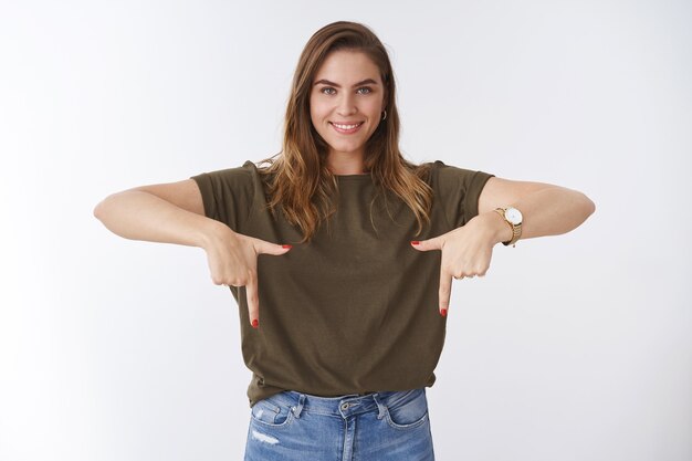 Studio shot attractive pleasant outgoing confident caucasian woman short hairstyle olive t-shirt pointing index fingers bottom down side smiling pleased showing promo recommending product