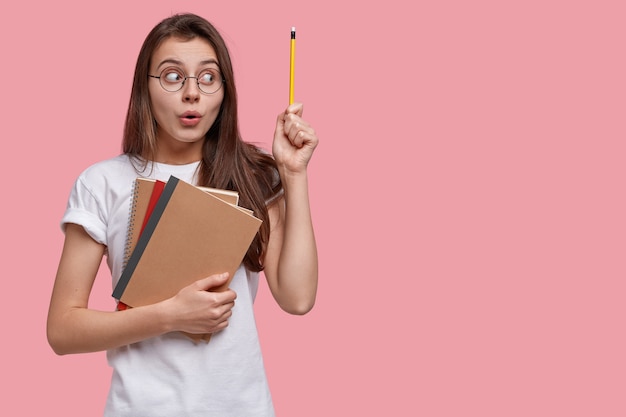 Studio shot of attractive woman has surprised expression, raises hand with pencil