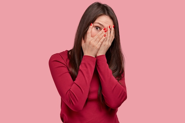 Studio shot of attractive woman looks through fingers, covers face with hands, has red manicure, wears casual clothes, poses against pink background