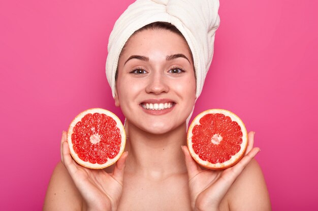 Studio shot of attractive woman with grapefruit in her hands and with white towel on her head, female after taking shower or bath, being in good mood, posing with toothy smile. Skin care concept.