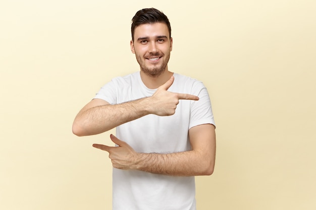 Studio shot di attraente giovane dai capelli scuri uomo in t-shirt bianca guardando la fotocamera con un ampio sorriso, puntando le dita anteriori in direzioni opposte, cercando di confonderti, mostrando il modo