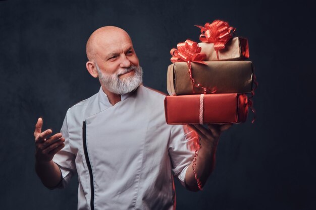 Studio shot of bald elderly chef dressed in uniform holding gift boxes.