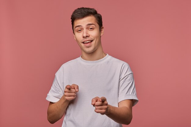 Studio shot of beautiful dark haired young male raising hand and pointing with index fingers ahead, standing over pink background in casual clothes, looking to camera self-confidently and smiling