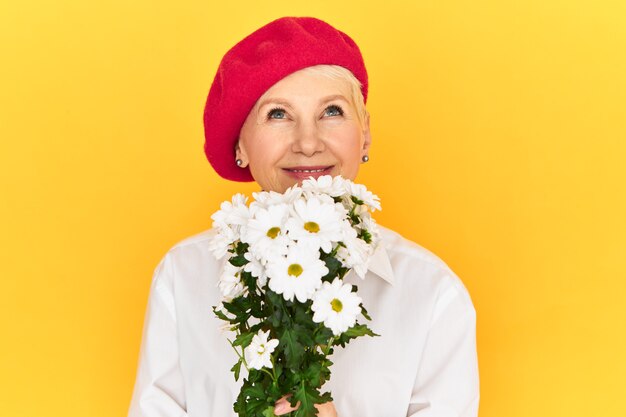 Studio shot of beautiful elegant French woman pensioner in stylish red beret enjoying aroma from white daisies, looking up with happy smile.