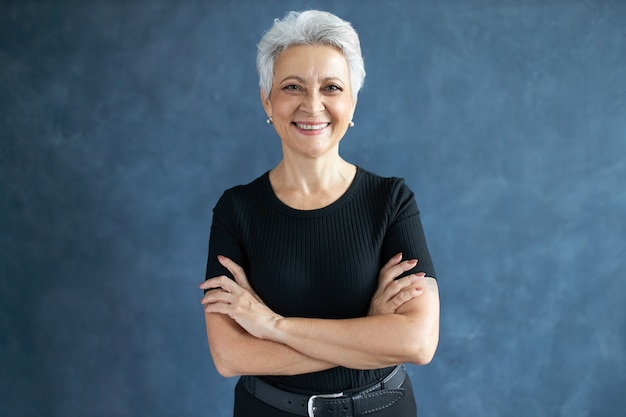 Studio shot of beautiful happy retired Caucasian female with pixie hairdo crossing arms on her chest, having confident look, smiling broadly