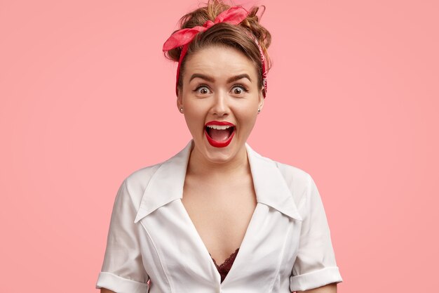 Free photo studio shot of beautiful joyful woman looks surprisingly at camera, opens eyes and mouth widely, feels astonished to recieve unexpected gift