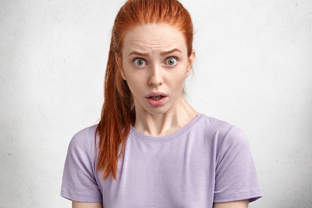 Studio shot of beautiful young woman with ginger hair and scared expression, stares at camera with bated breath, being dissatisfied