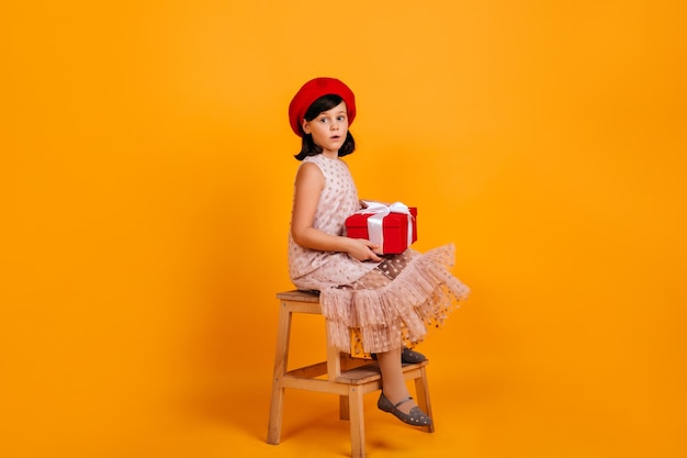 Studio shot of birthday girl sitting on chair Female child in french beret posing with gift on yellow background