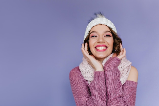 Studio shot of blissful dark-haired girl with pink lips. Indoor portrait of carefree curly female model in woolen outfit posing on purple background.