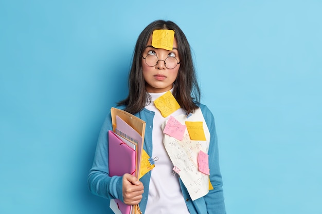 Studio shot of brunette young Asian schoolgirl studies mathematics concentrated above on forehead with sticker holds folders