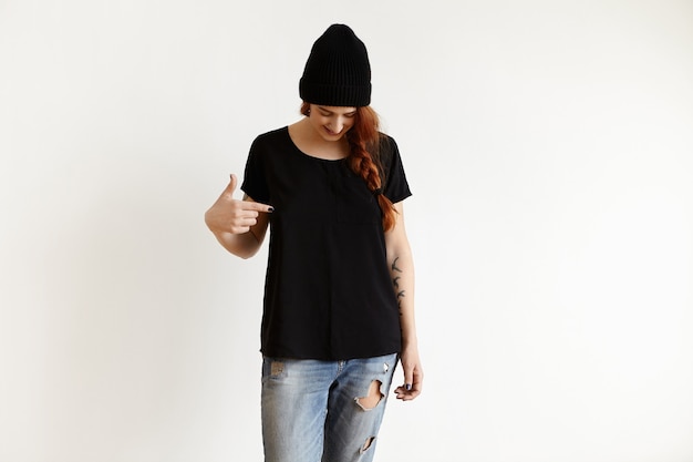 Studio shot of Caucasian girl in stylish hat and ragged blue jeans, looking down and pointing index finger