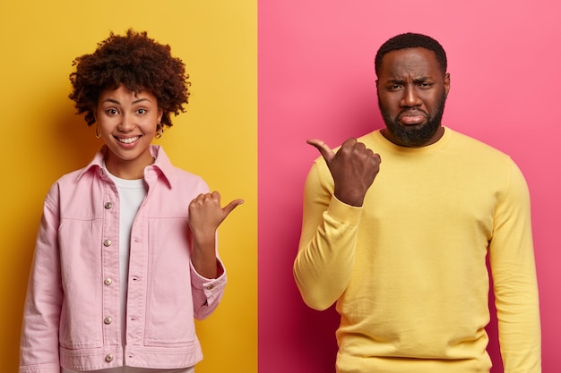 Studio shot of cheerful black woman and gloomy bearded man point thumbs at each other, blame and express different emotions