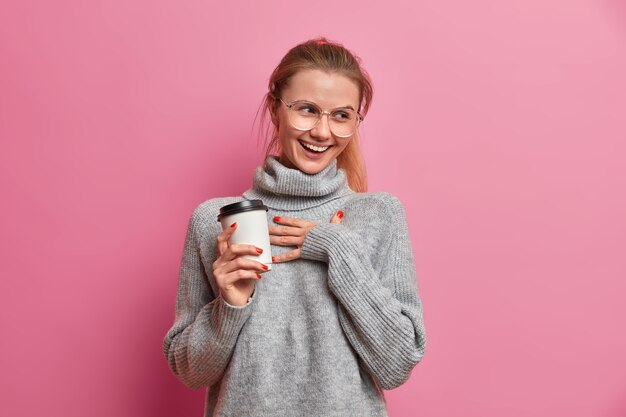 Studio shot of cheerful European girl giggles positively keeps hand on chest and holds takeaway coffee