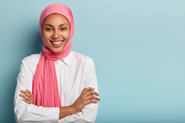 쾌활한 종교 무슬림 여성의 스튜디오 샷은 팔을 접고 넓게 미소 짓고 하얀 치아를 가지고 있습니다.