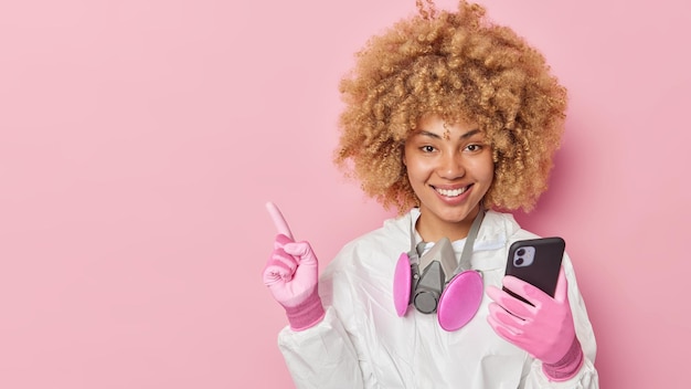 Studio shot of cheerful young woman ecologist wears protective white suit and gloves works in toxin area holds mobile phone prevents dangerious ecological problems isolated over pink background