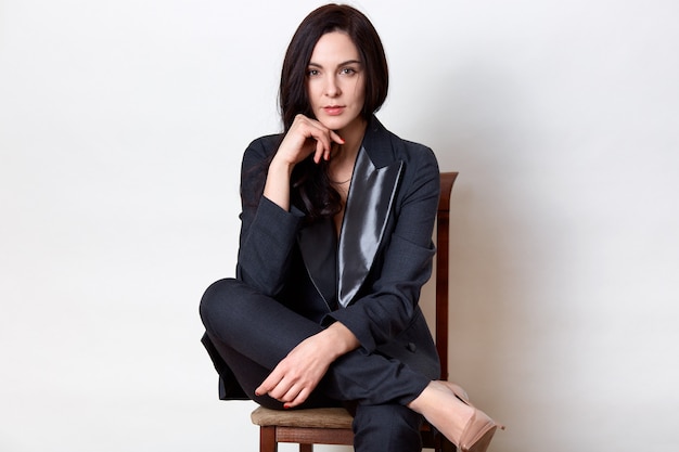 Studio shot of confident brunette woman sitting on wooden chair and keeping fist under chin