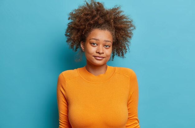 Studio shot of confused dark skinned teenage girl has curly bushy hair shrugs shoulders feels doubt wears casual orange long sleeved jumper