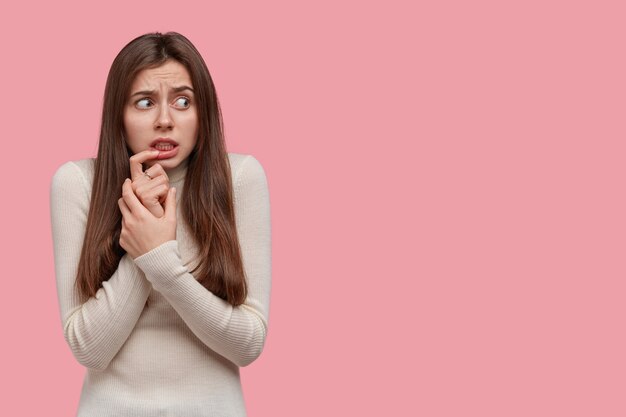 Studio shot of confused nervous woman keeps index finger on lips, looks with scared expression aside, dressed in white jumper