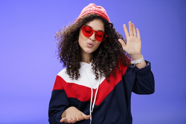 Studio shot di una ragazza dj cool ed elegante in berretto rosso e occhiali da sole che alza il palmo mentre si gode musica fresca e balla al ritmo delle labbra pieganti divertendosi alla fantastica festa in posa sul muro blu.