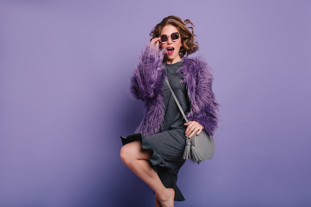 Studio shot of curly barefooted girl in fur coat jumping during photoshoot