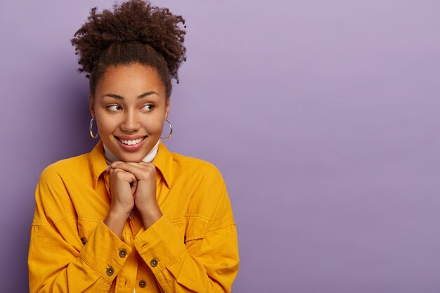 Studio shot of dark skinned woman with curly combed hair, wears earrings and denim yellow jacket, keeps hands under chin, looks aside, expresses sincere emotions, smiles carefree, copy space aside