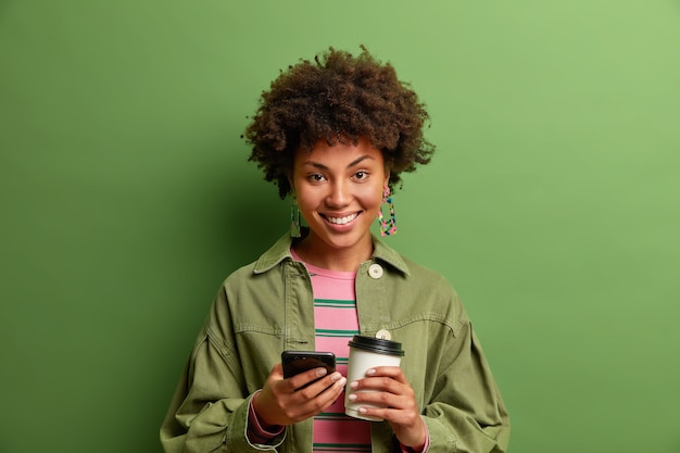Free photo studio shot of dark skinned young woman holds mobile phone and take away coffee