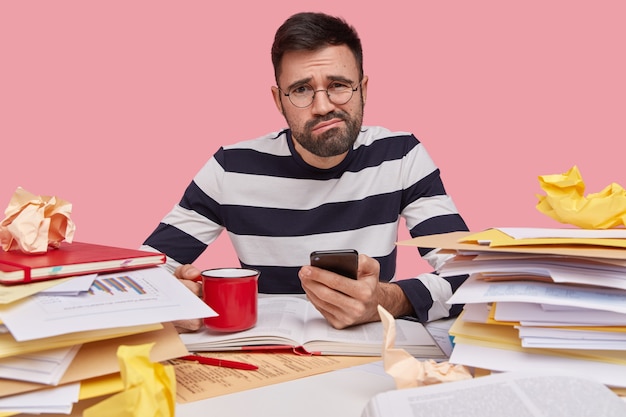 Studio shot of displeased Caucasian man frowns face, holds cellular in hand, recieves message with tasks to do, wears striped jumper, drinks hot coffee or tea