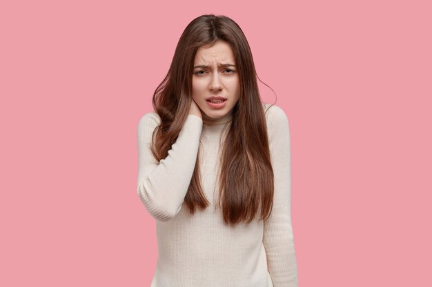 Studio shot of displeased European woman frowns face in discontent, keeps hand on neck, cries from pain, wears casual jumper, models over pink studio wall. People