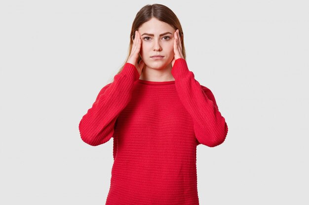 Studio shot of displeased female suffers from headache, dressed in red sweater, keeps hands on temples, has upset facial expression