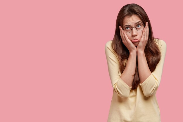 Free photo studio shot of displeased upset woman purses lips, keeps hands on cheeks, dressed in casual yellow sweater