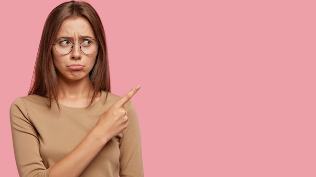 Studio shot of displeased woman purses lips, points with index finger at upper right corner, has unhappy expression