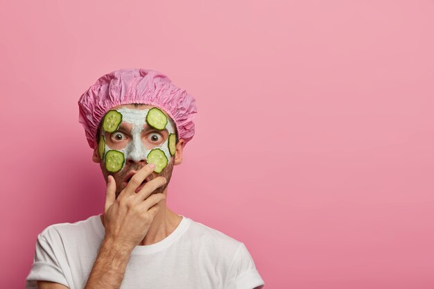 Studio shot of emotional European man covers mouth with palm, shocked to hear information from beautician, applies cucumbers and clay mask on face