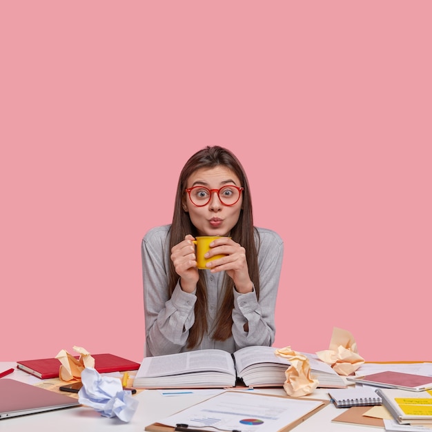 Studio shot di emotiva bella donna con lunghi capelli lisci, fa smorfia, beve il tè, si prepara per la lezione