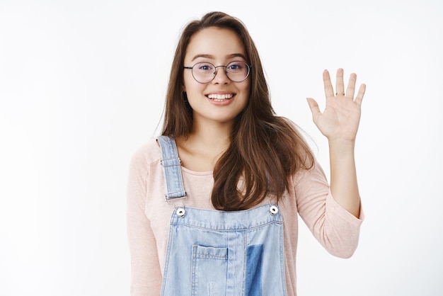 興奮した魅力的なフレンドリーな女の子が、カメラを見つめているハイジェスチャーで手のひらを上げてこんにちはを振って、肩を不器用に上げて驚いて広く笑っている新しい人々に会うスタジオショット