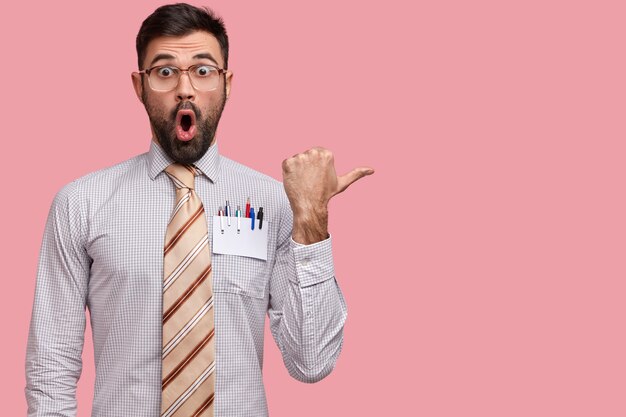 Studio shot of experienced male employee with widely opened mouth, feels surprisement and astonishment, wears elegant shirt, points aside at blank space