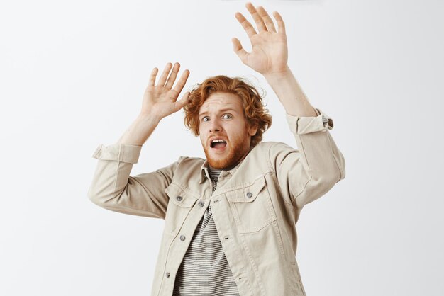 Studio shot of frightened timid redhead guy was scared by friend raising palms in surrender