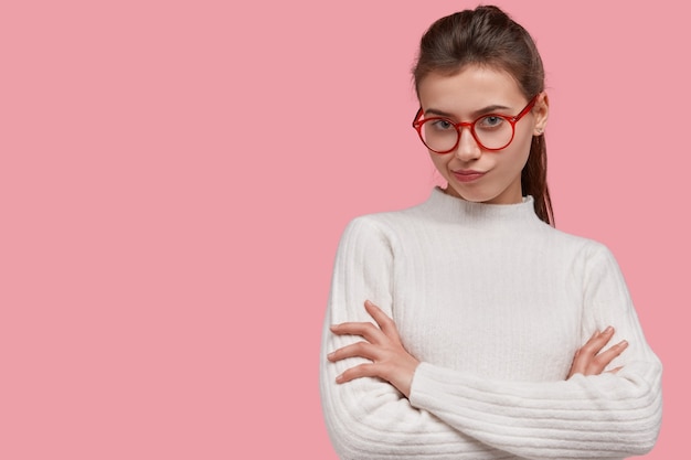 Studio shot of frustrated confused woman keeps arms folded, purses lips, wears optical glasses