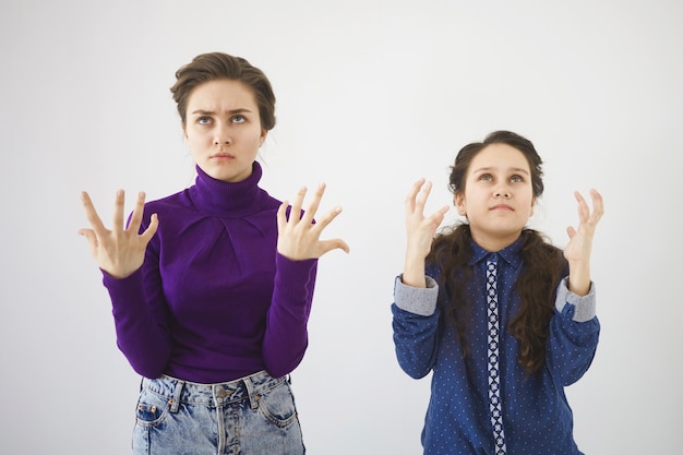 Студийный снимок разъяренной раздраженной девочки-подростка и ее сестры, позирующих у белой стены, эмоционально жестикулирующих и сердито смотрящих вверх
