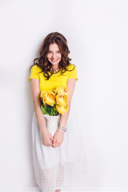 A studio shot of a girl standing and holding a white vase with flowers. The girl has long wavy brunette hair and wears a yellow t-shirt and white skirt. Girl smiles widely and looks at the camera.