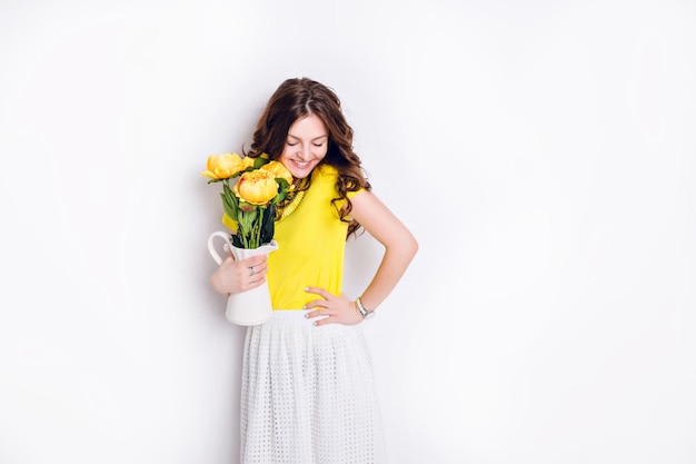 Студийный снимок девушки, стоящей и держащей белую вазу с цветами. У девушки длинные волнистые волосы брюнетки, она носит желтую футболку и белую юбку. Девушка широко улыбается.