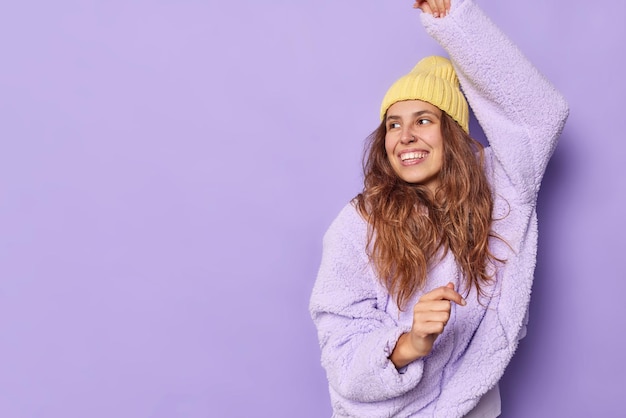 Free photo studio shot of glad european female model shakes arms looks happily dressed in warm outerwear has piercing in nose dances carefree isolated over purple background with copy space on left side