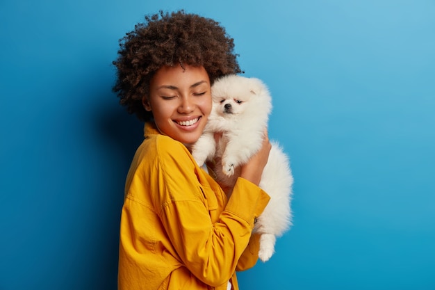 Free photo studio shot of glad female model holds fluffy pet closely to face after grooming, smiles with happiness