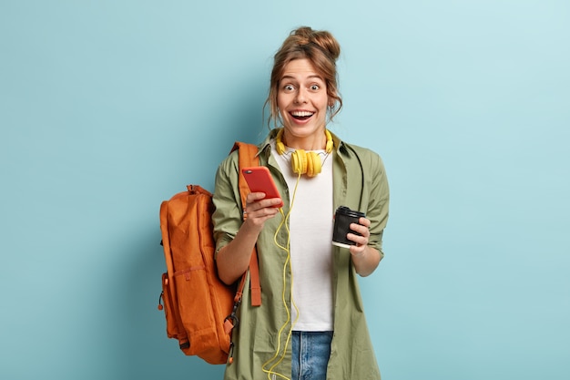 Studio shot of glad hipster girl holds modern cell phone, checks notification, connected to stereo headphones